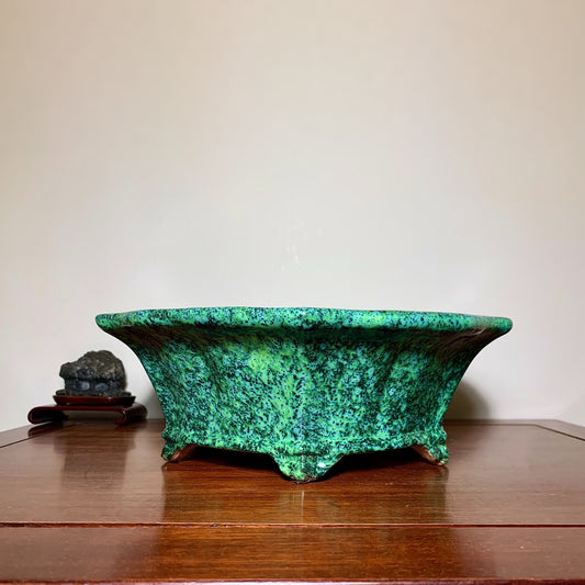 A wide, shallow green glazed pot with speckled texture and three feet, sits on a wooden surface.
