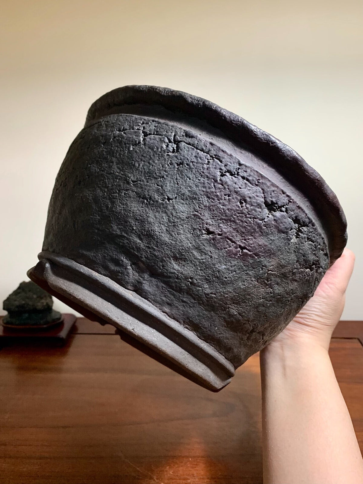 A hand holds a dark, rough-textured, unglazed ceramic pot with a wide rim and visible cracks.