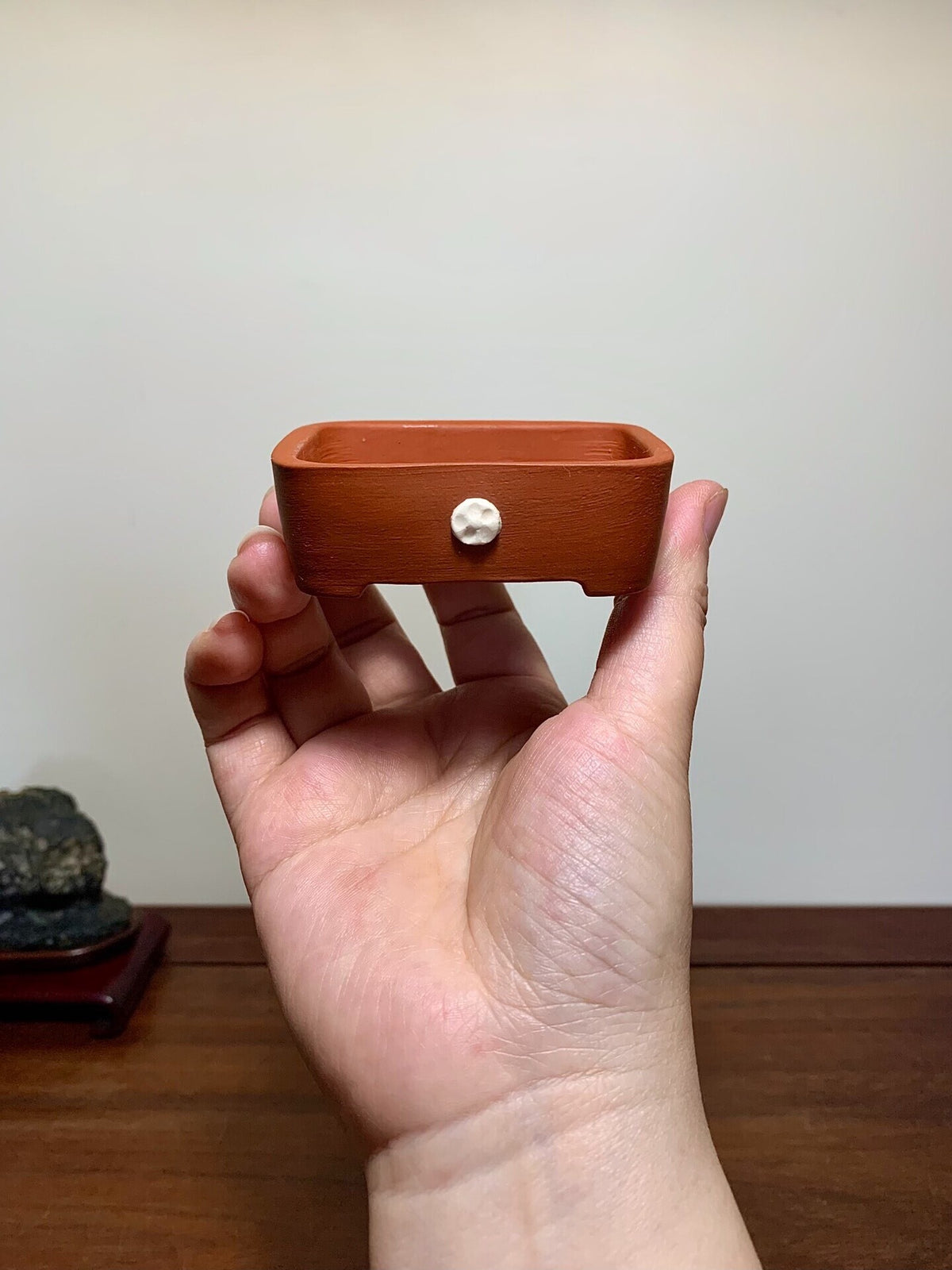A hand holds a small, unglazed terracotta rectangular pot with a single white knob on its side.