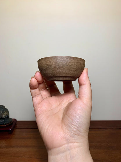 A hand holds a small, brown, matte-finished ceramic bowl with a wide rim and gently sloping sides.