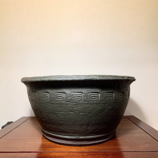 Dark grey unglazed ceramic pot with repeating geometric relief pattern around its midsection.