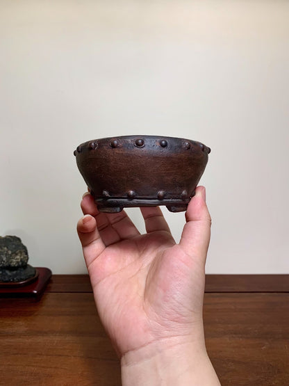 A hand holds a small, dark brown, unglazed ceramic pot with raised bumps around the rim.