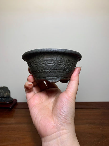 A hand holds a dark grey, round, unglazed ceramic pot with raised geometric patterns.