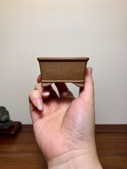 A small, unglazed brown square pot with a flat rim and four short feet, held in a hand.