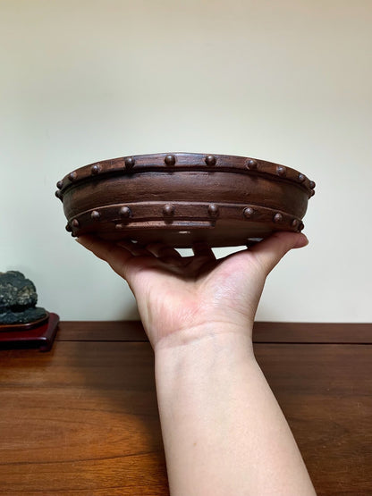 A hand holds a dark brown, round, unglazed ceramic pot with raised bumps around its rim.