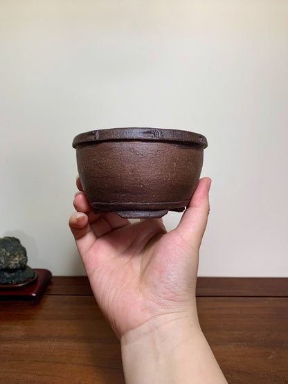A hand holds a small, dark brown, unglazed ceramic pot with a textured surface and a slightly flared rim.