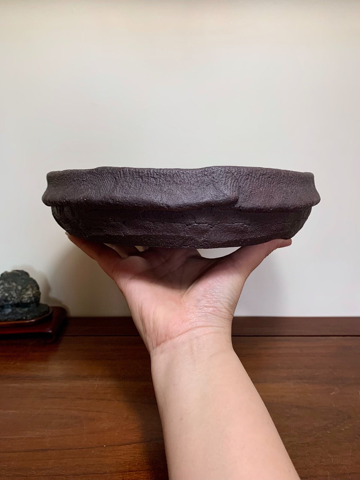 A hand holds a dark brown, textured, unglazed ceramic pot with a wide, shallow bowl and uneven rim.