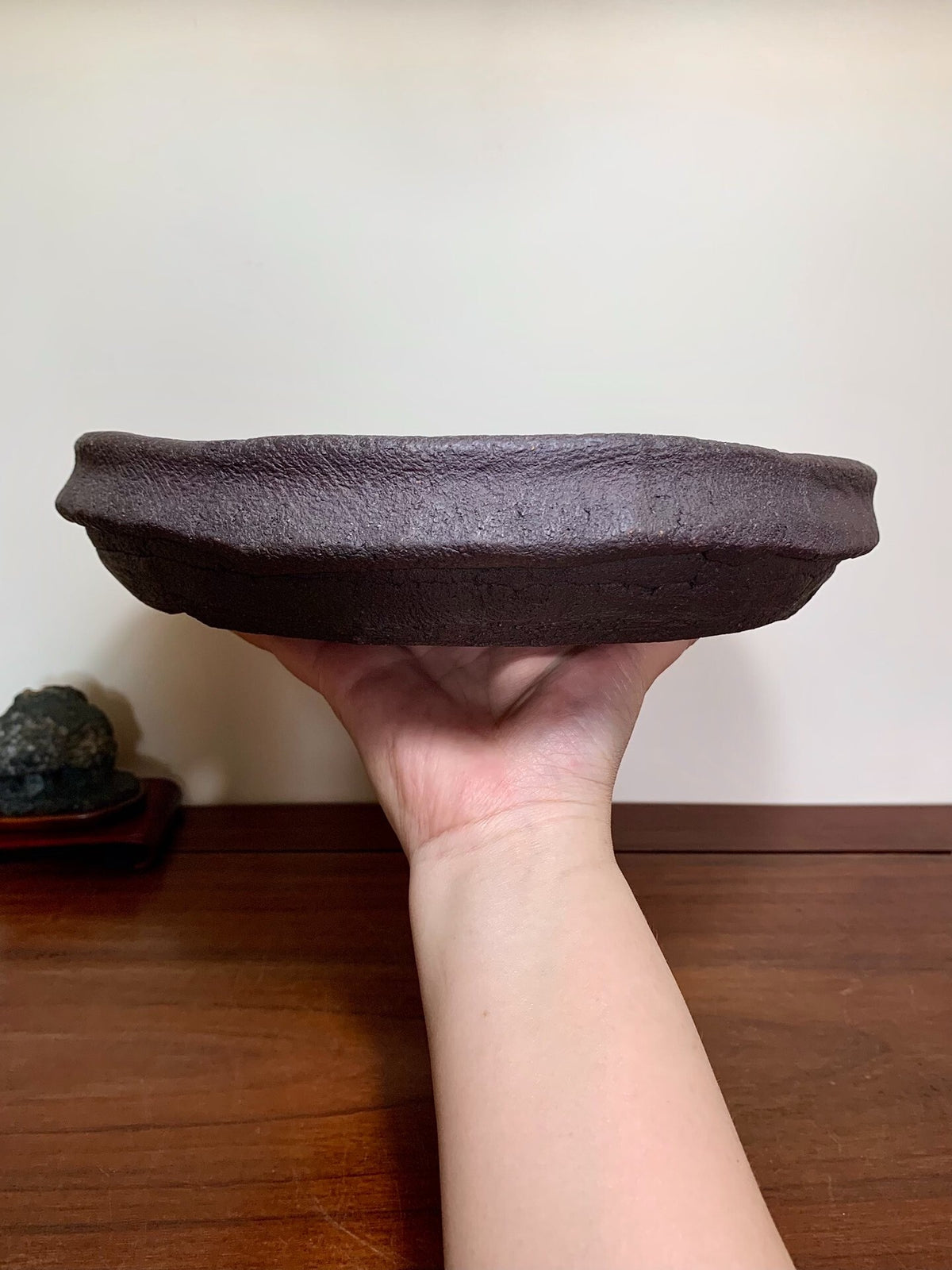 A hand holds a dark, textured, unglazed ceramic pot with a wide, shallow rim.