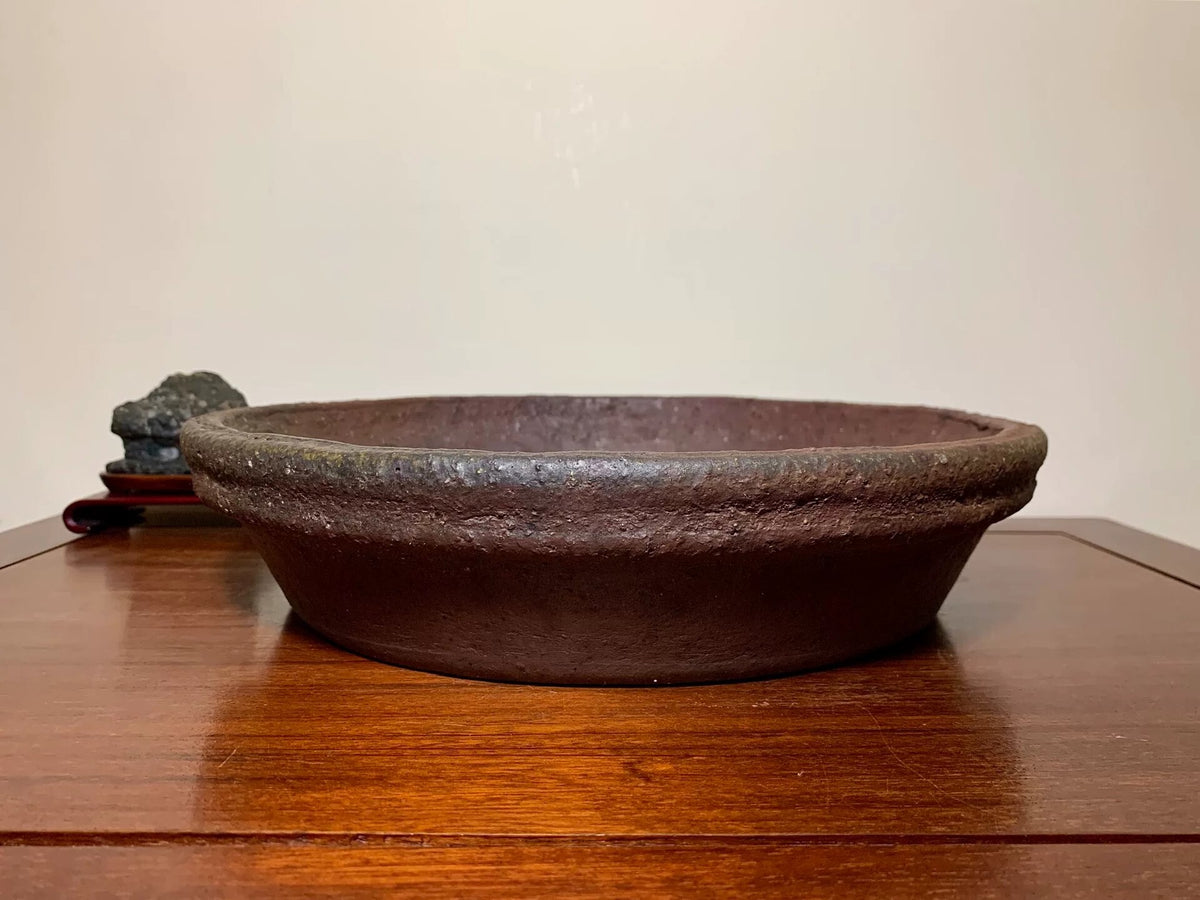 Dark brown, shallow, unglazed ceramic pot with rough texture and slightly raised rim.