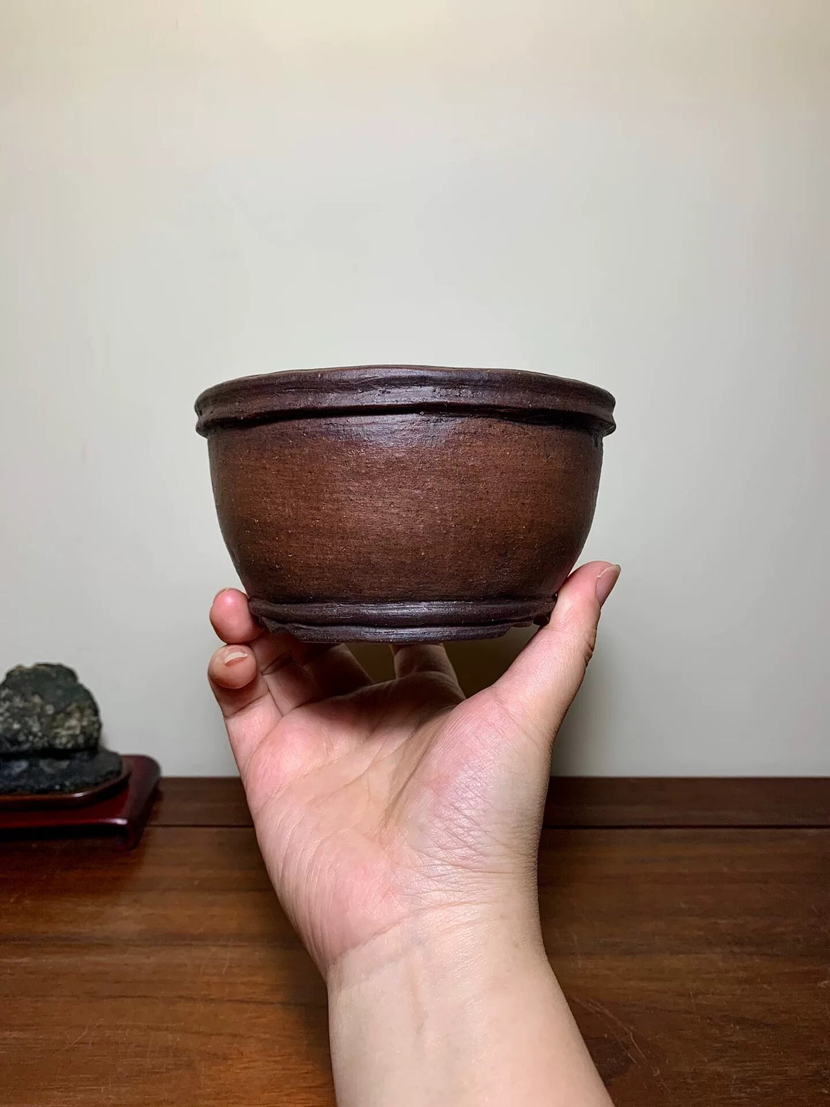 A hand holds a small, dark brown, unglazed ceramic pot with a wide rim and rounded body.
