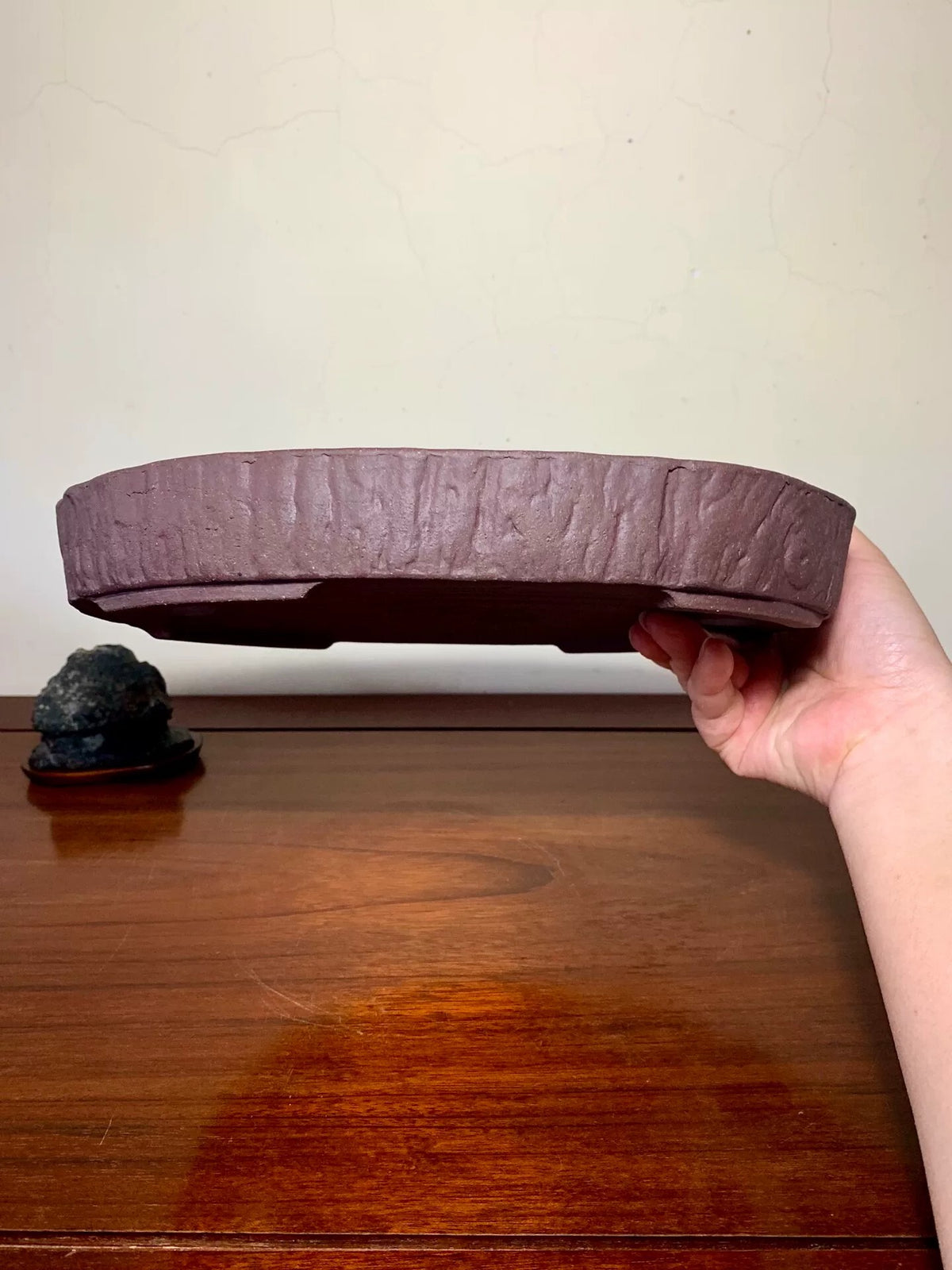A hand holds a dark brown, textured, unglazed ceramic pot with a wide, shallow, oval shape.