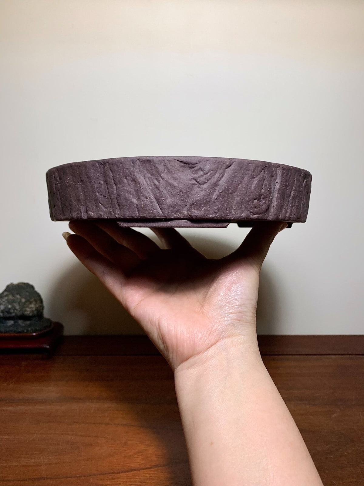 A hand holds a dark brown, round, unglazed pot with a textured, tree-ring-like surface.