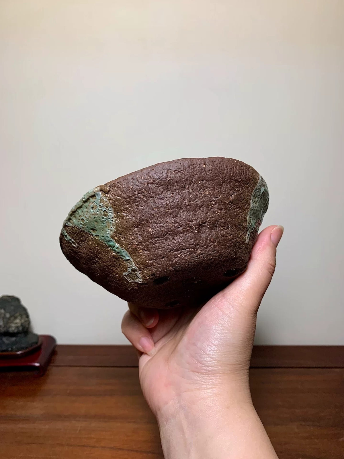A hand holds a rough, brown, irregularly shaped stone with greenish patina patches.