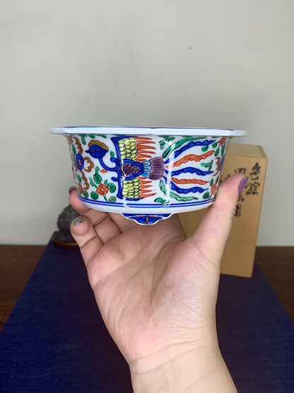 A hand holds a small, white glazed ceramic pot with colorful painted birds and foliage.