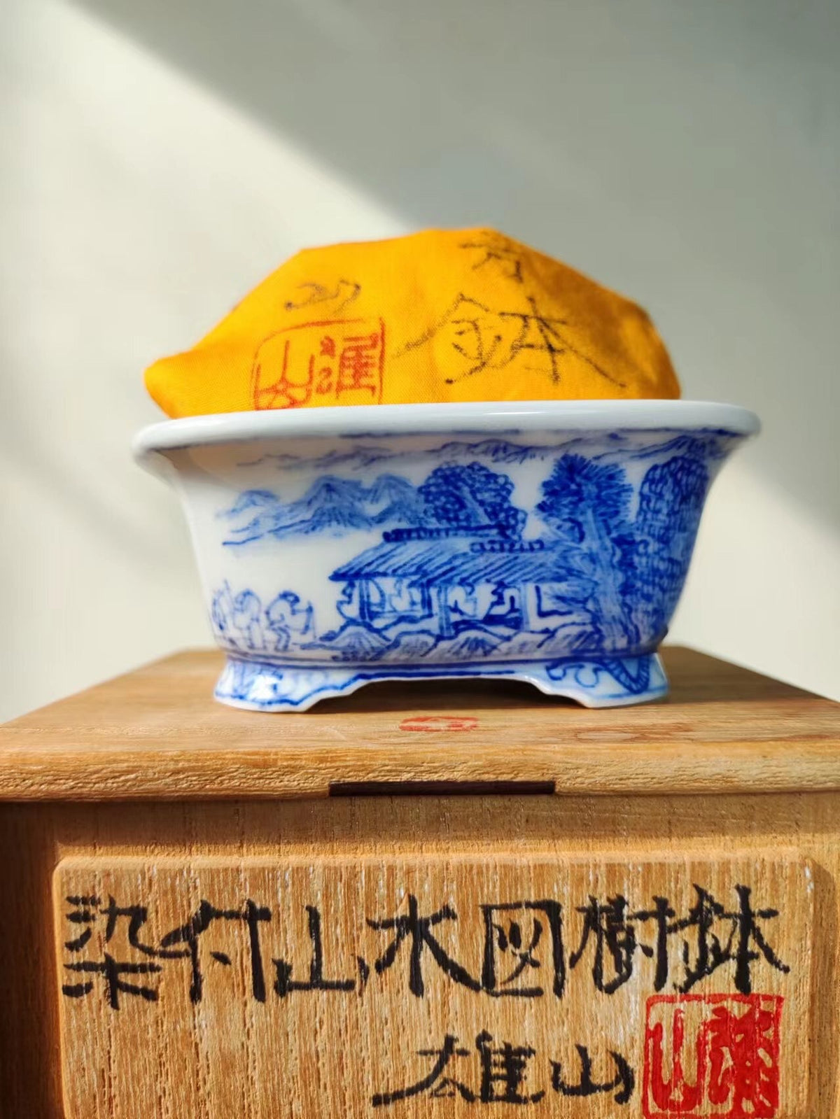 Blue and white ceramic pot with mountain scene, covered by yellow cloth with red seal.