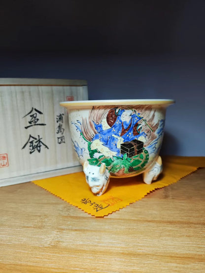 A white ceramic pot with blue, green, and brown painted figures on three animal-shaped feet.