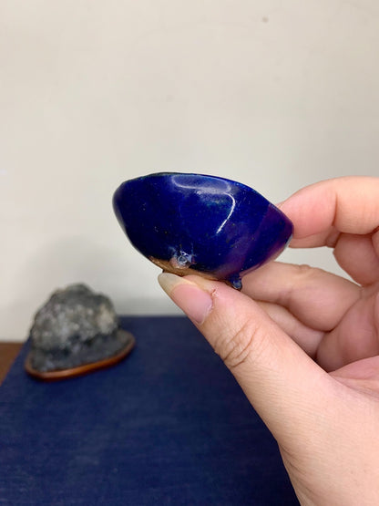 A hand holds a glossy, deep blue, rounded pot with a brown rim and a small white mark.