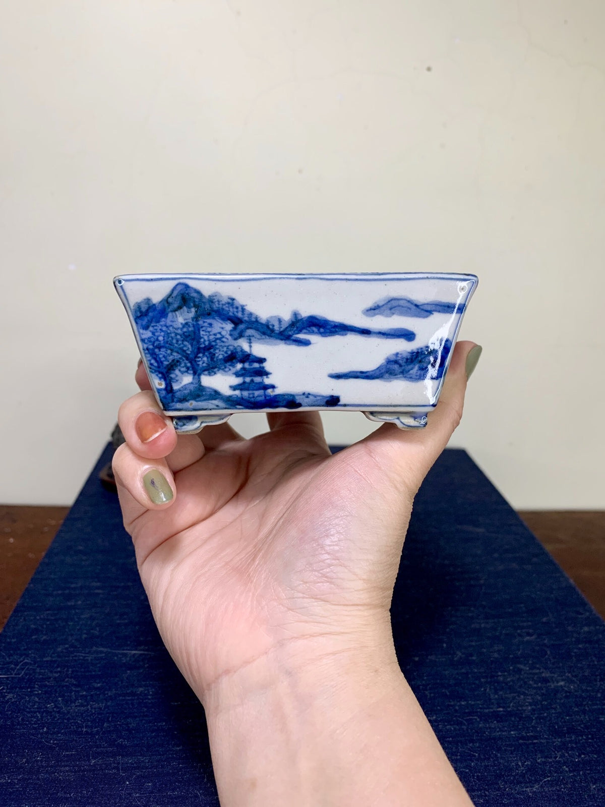 A hand holds a small, rectangular blue and white glazed pot with painted mountain scenery.