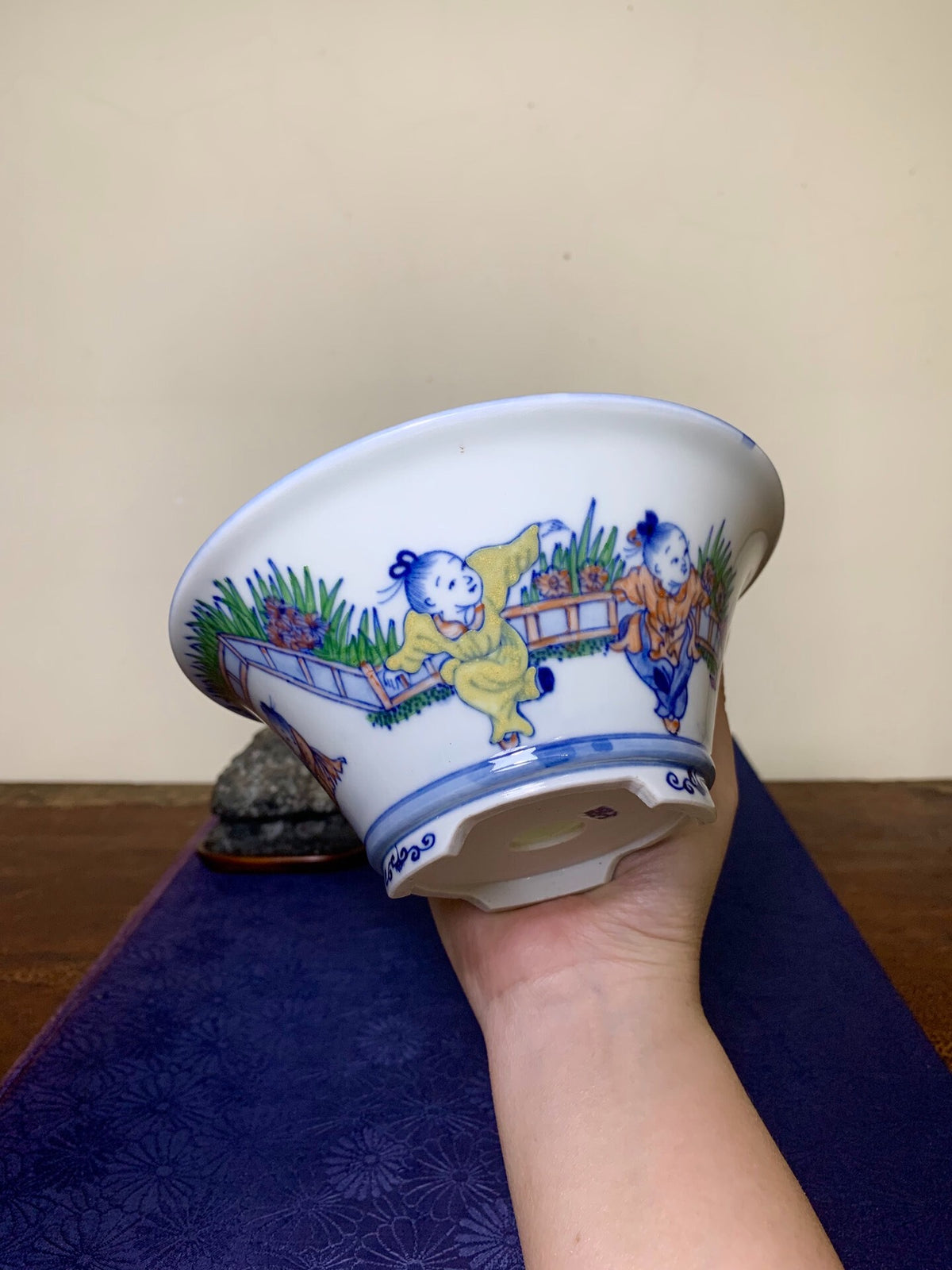 A hand holds a white ceramic pot with blue and yellow painted children on a bridge, glossy finish.