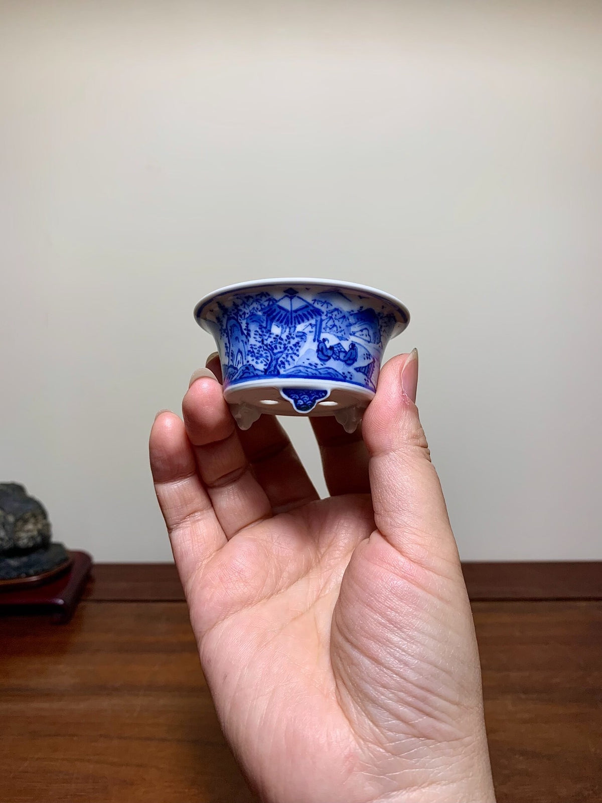 A hand holds a small blue and white ceramic pot with a landscape design.