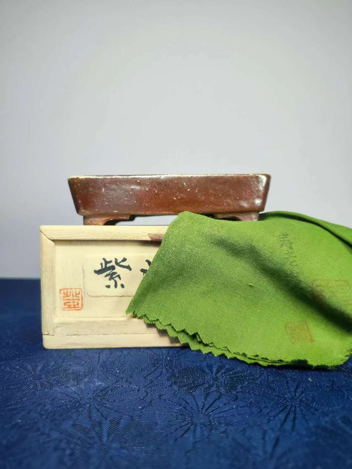 A brown glazed rectangular pot rests atop a light wooden box with black characters and red seal, beside a green cloth.