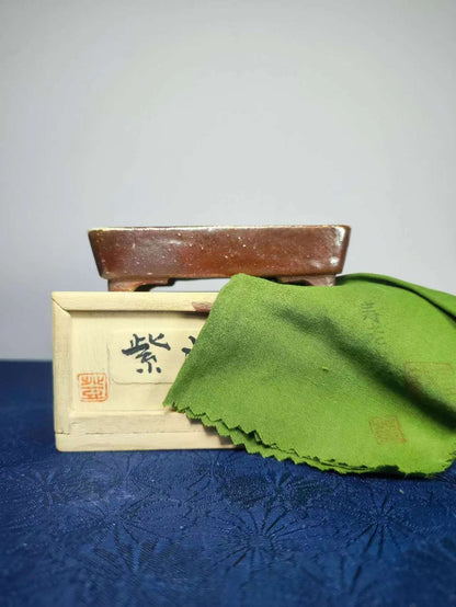 A brown glazed rectangular pot rests atop a light wooden box with black characters and red seal, beside a green cloth.