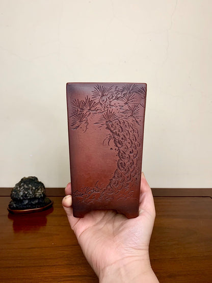 A hand holds a square, reddish-brown glazed pot with an engraved pine tree design.