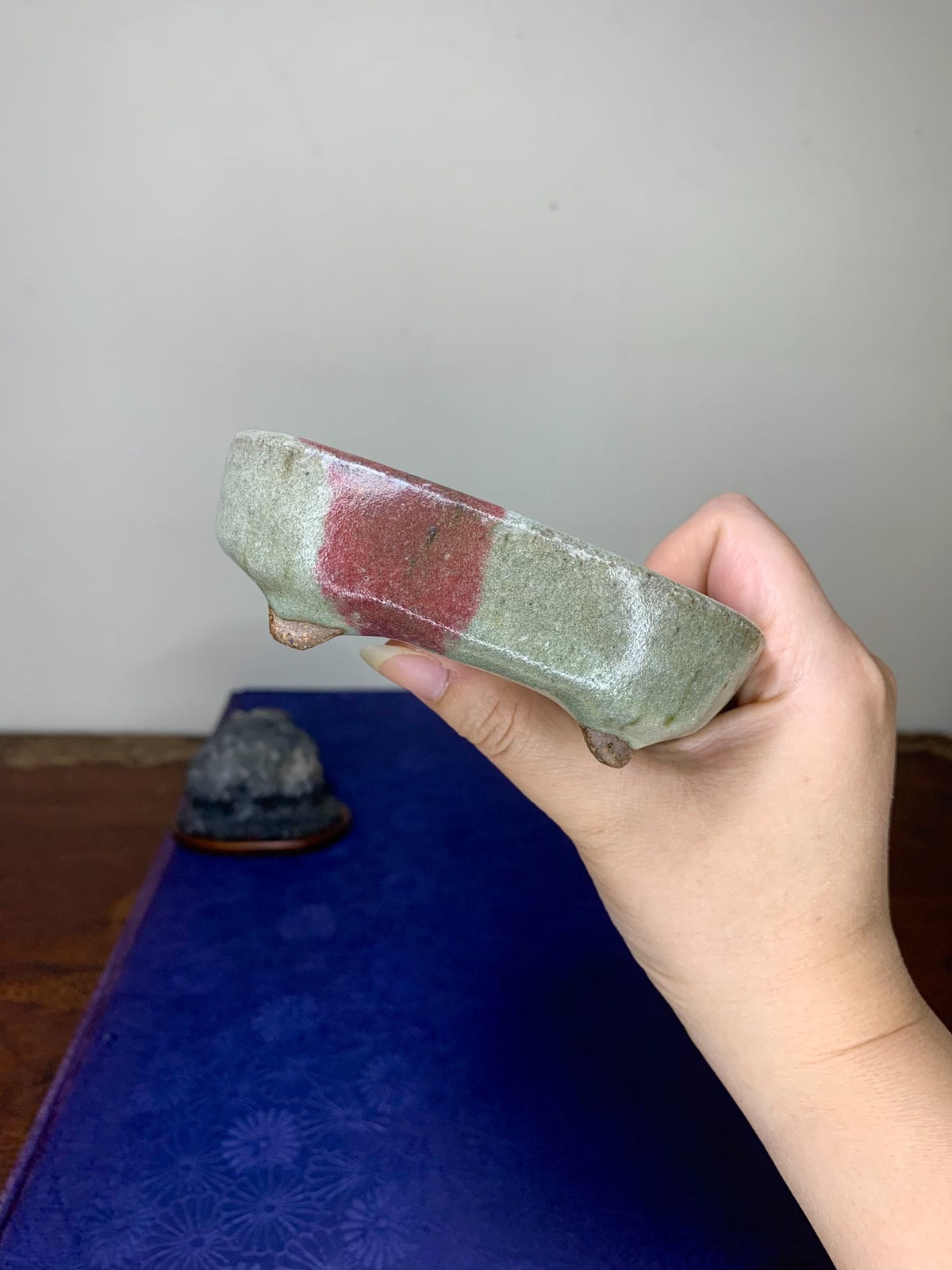 A hand holds a small, rectangular glazed pot with mottled green and red glaze.