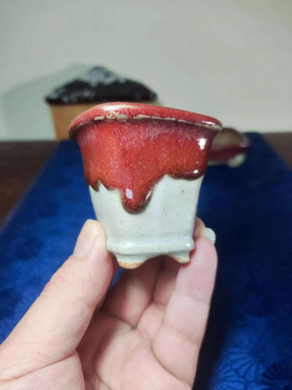 A hand holds a small ceramic pot with a glossy red glaze dripping down its sides over a matte grey base.