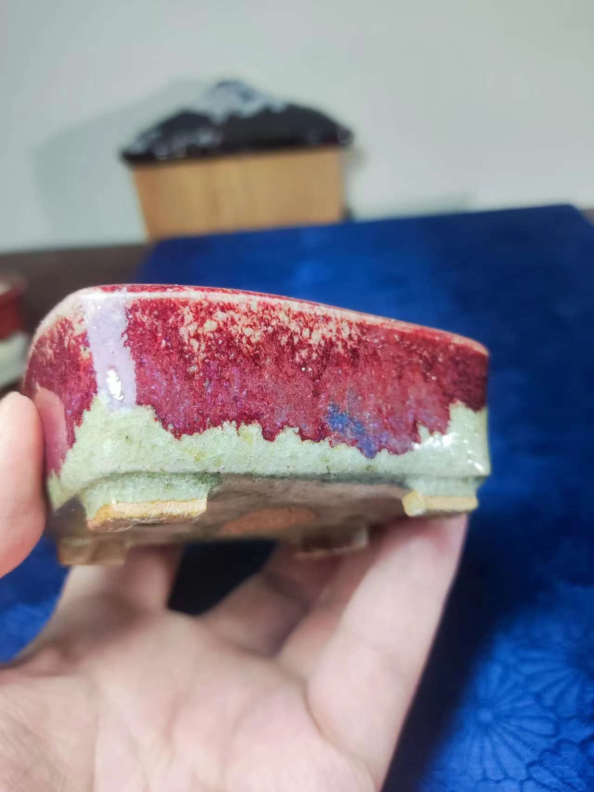 A hand holds a small, rectangular ceramic pot with a glossy red and green glaze.