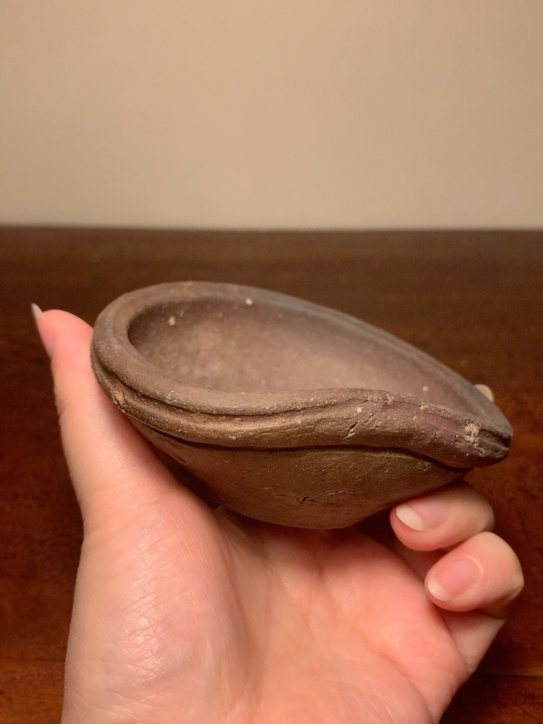 A small, unglazed brown clay pot with a shallow, rounded bowl held in a hand.
