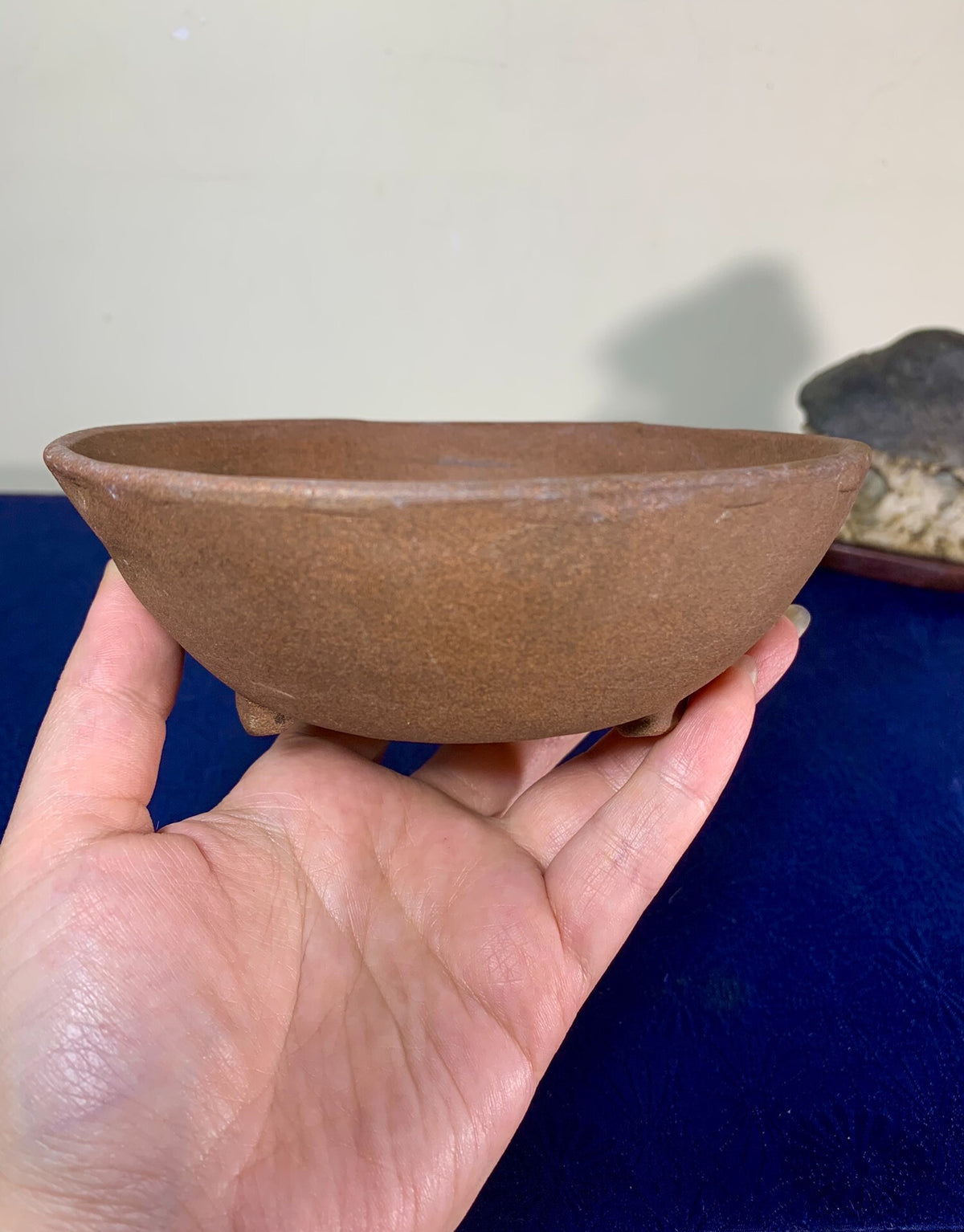 A hand holds a small, unglazed brown ceramic pot with a wide rim and three feet.