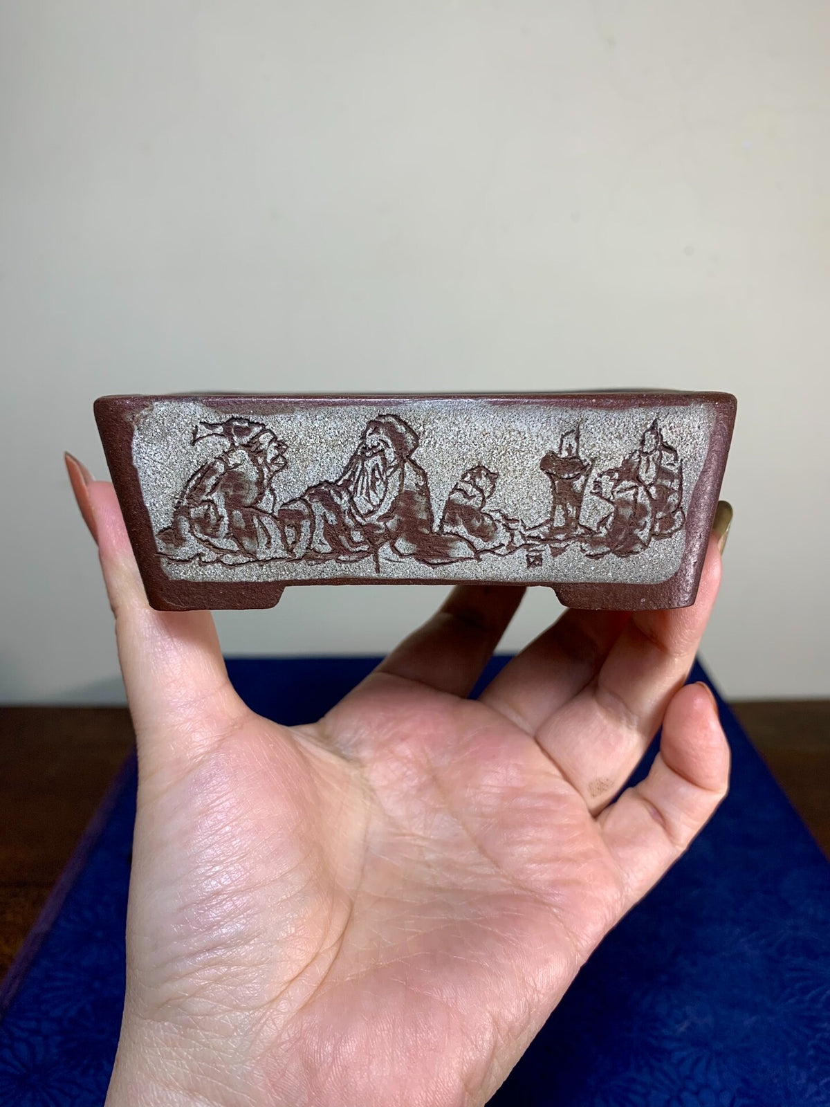 A hand holds a rectangular, matte brown ceramic pot with a grey, textured surface and incised figures.