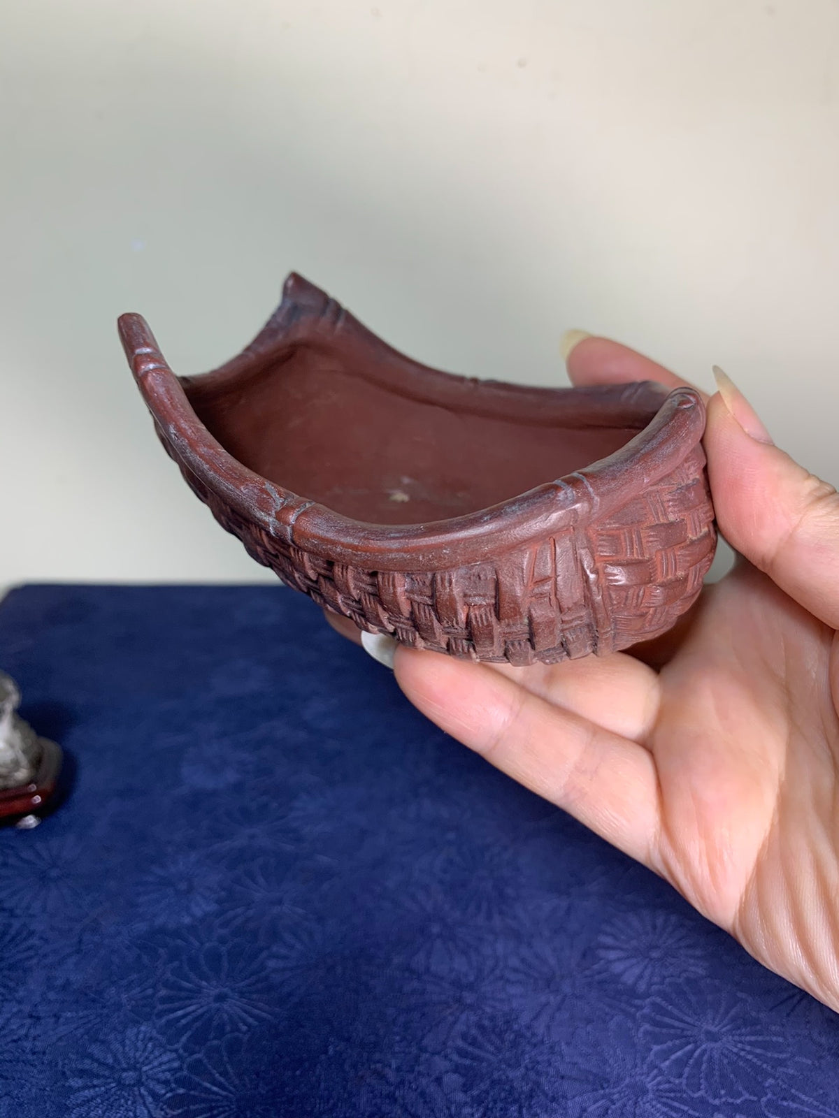 A hand holds a reddish-brown, curved ceramic pot with a woven pattern relief.