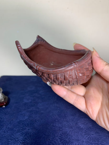 A hand holds a reddish-brown, curved ceramic pot with a woven pattern relief.