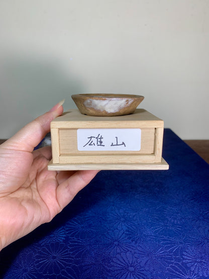 Hand holds small wooden stand with label and a shallow brown glazed ceramic bowl on top.