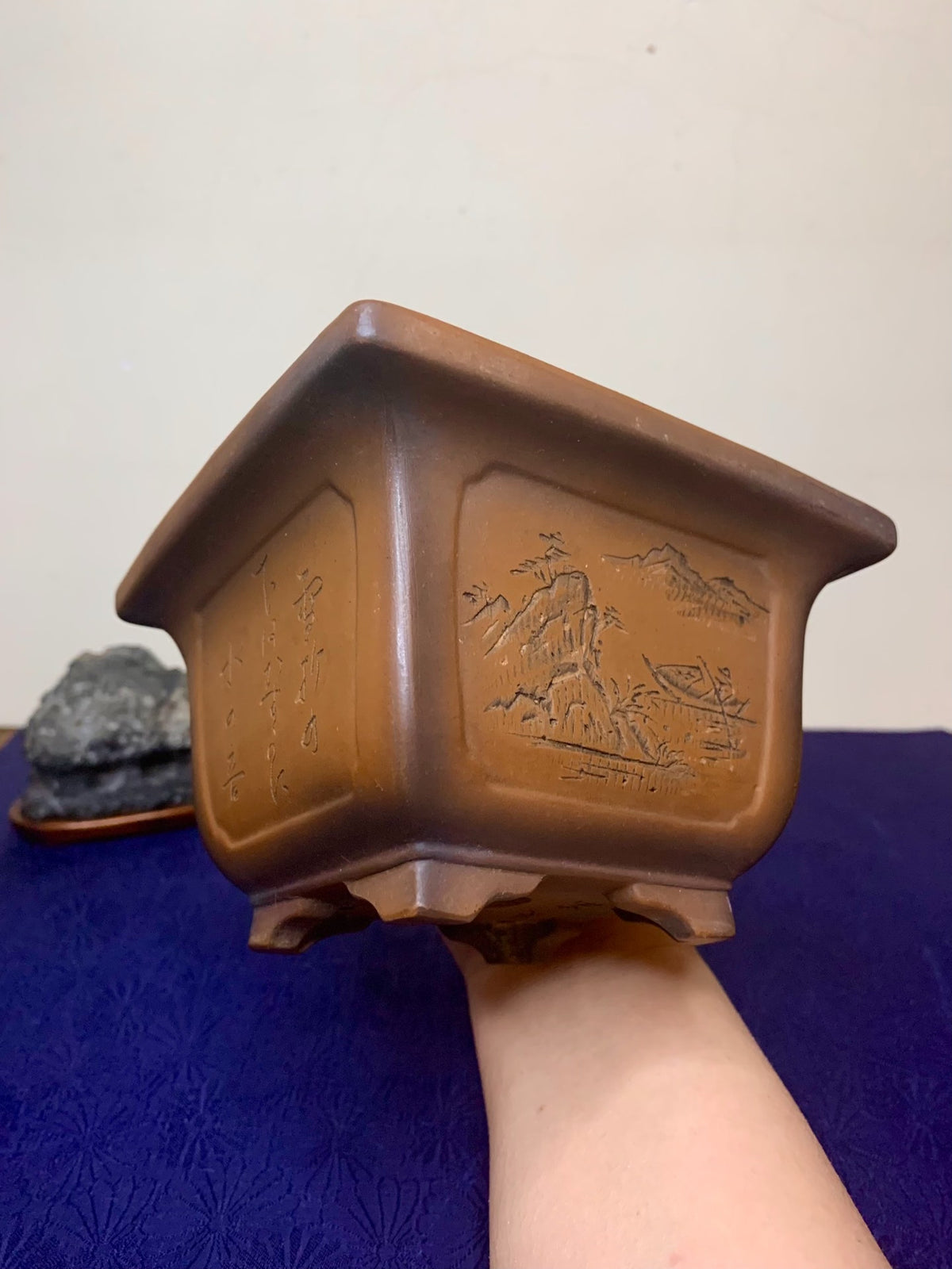 A brown, square ceramic pot with carved mountain and boat scenes and vertical script on its sides.