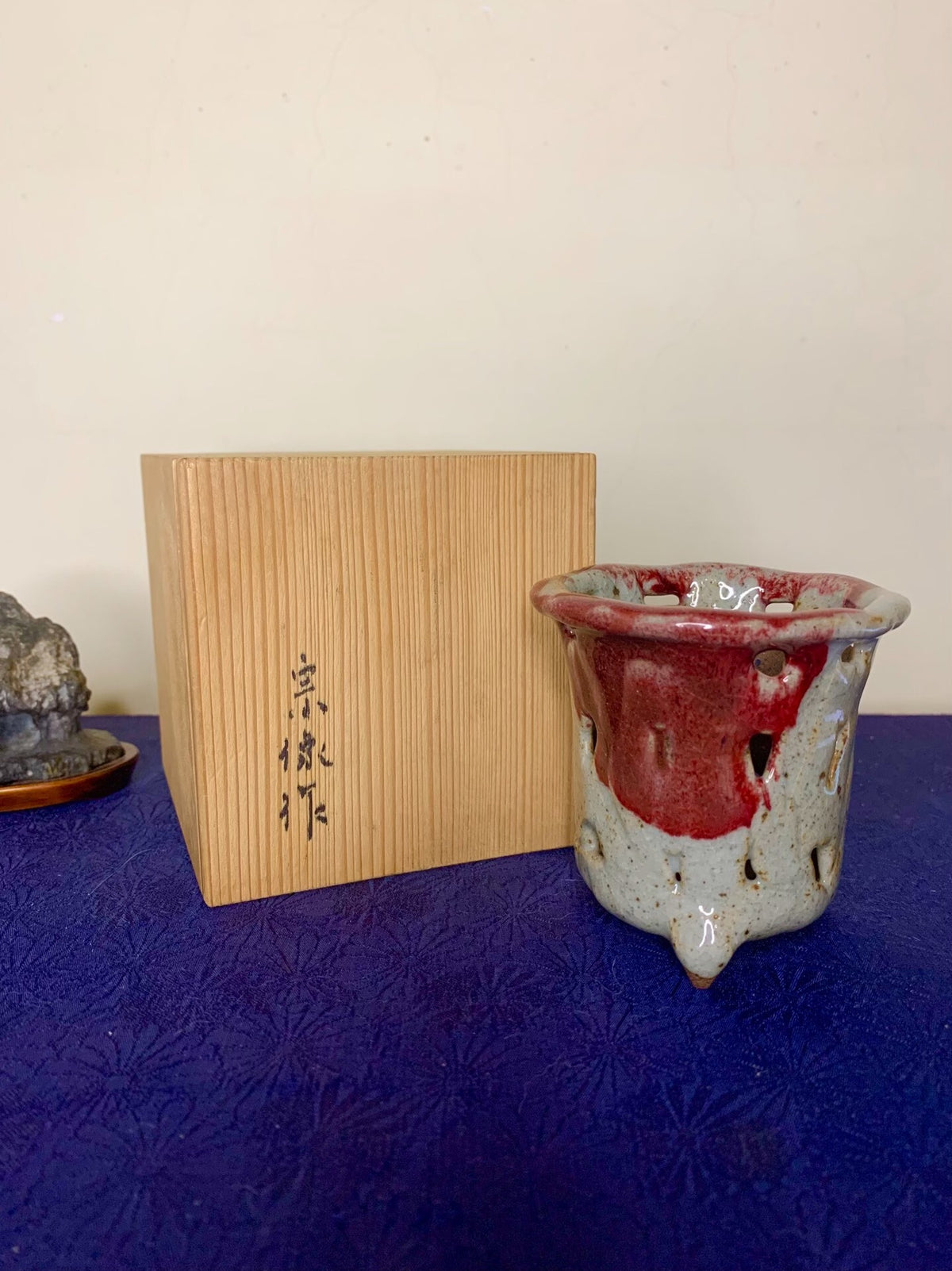 A small, glazed ceramic pot with red and grey glaze, three feet, and holes around its rim.