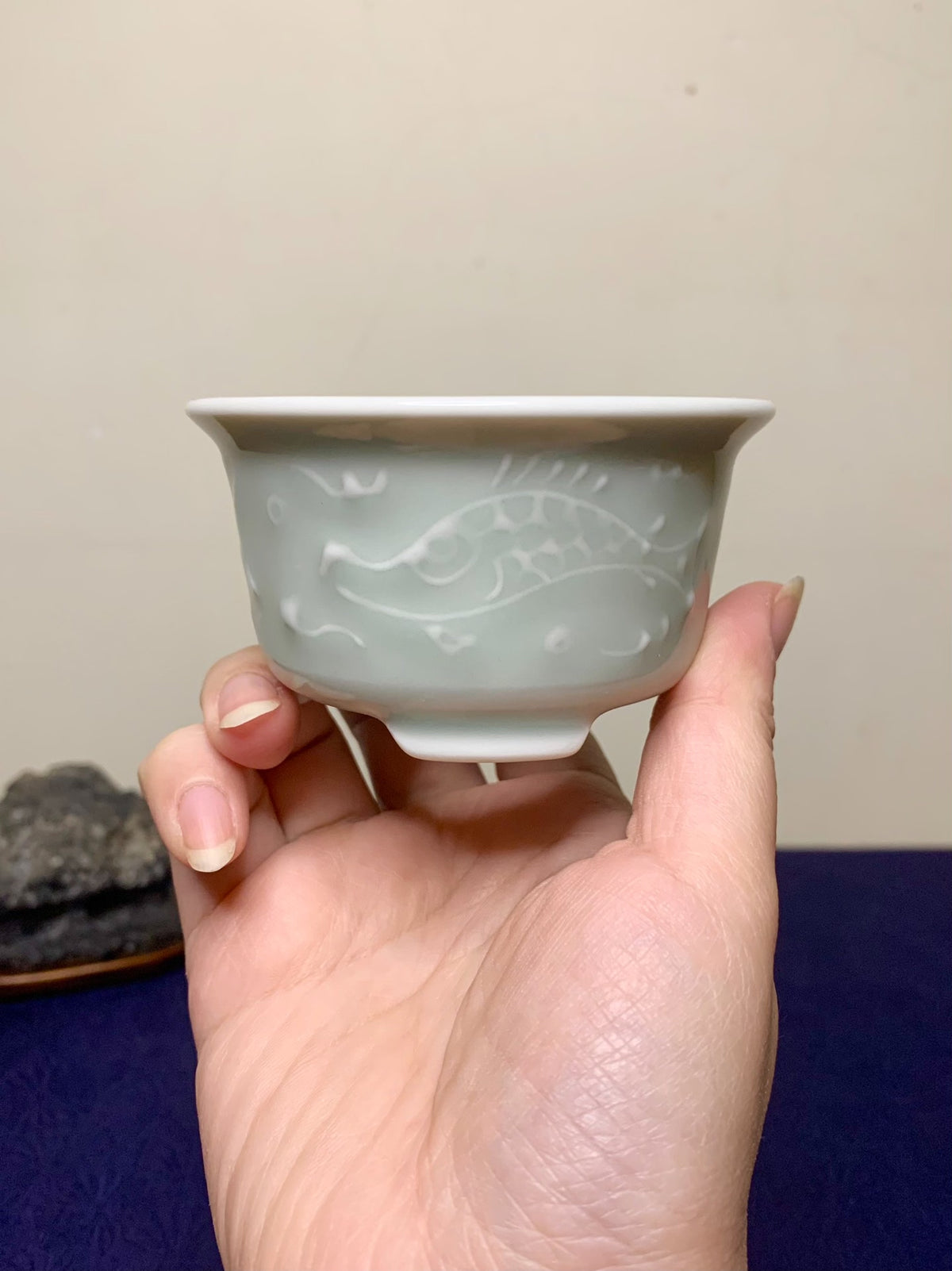 A hand holds a pale green glazed ceramic cup with an embossed fish design.