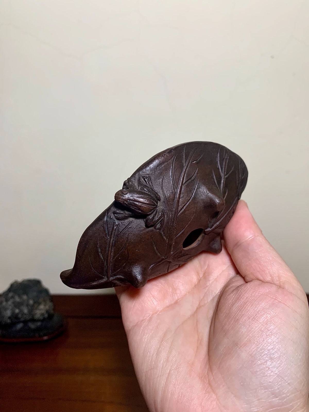 A hand holds a dark brown, leaf-shaped carved object with a hole and twig details.