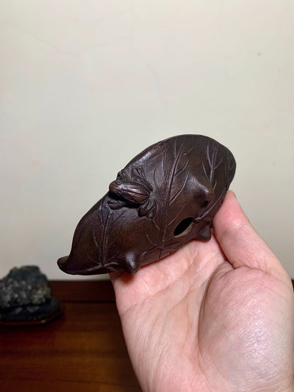 A hand holds a dark brown, leaf-shaped carved object with a hole and twig details.