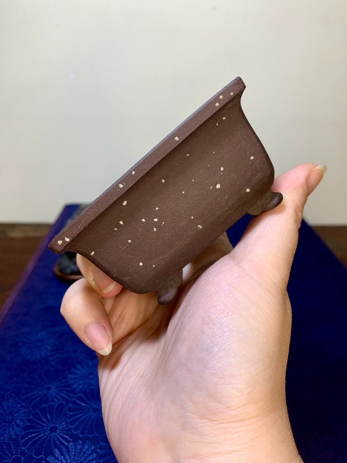 A hand holds a small, brown, unglazed rectangular pot with scattered light speckles.