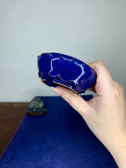 A hand holds a small, deep blue glazed pot with white crackle glaze and rivet-like dots.