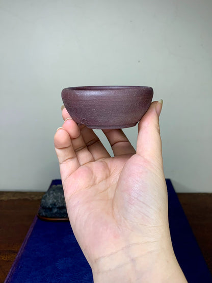 A hand holds a small, matte, deep purple unglazed ceramic bowl with a rounded rim.