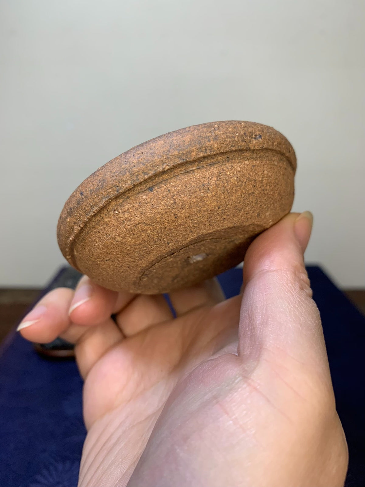 A hand holds a small, round, unglazed brown ceramic pot base with a textured surface and a central indentation.