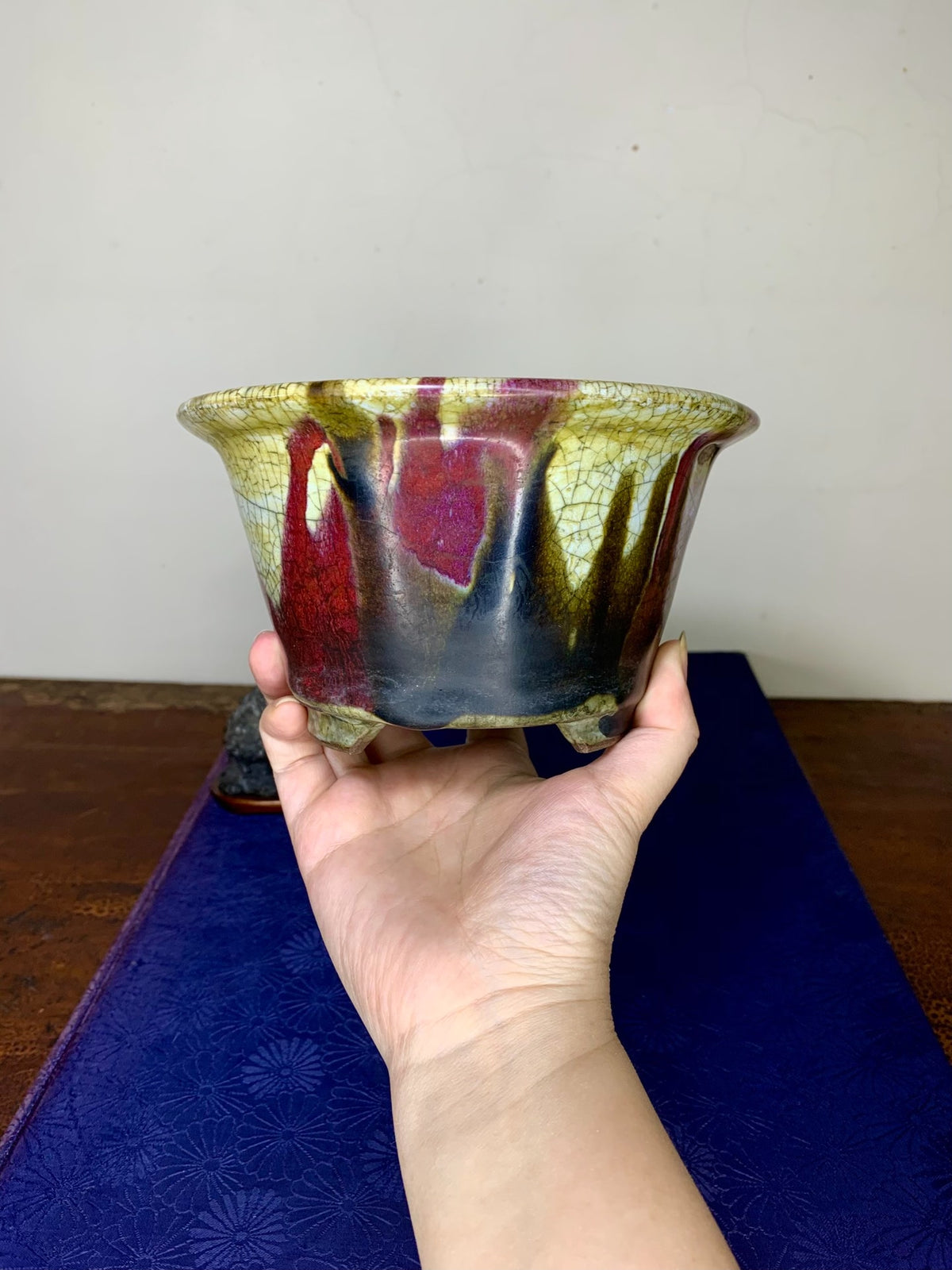 A hand holds a small, three-footed ceramic pot with a crackled, multicolored glaze.