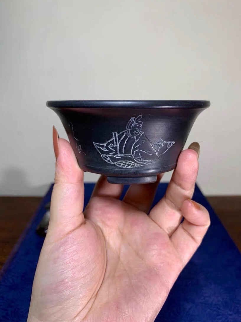 A hand holds a small, dark, glossy bowl with a white etched figure of a seated person.