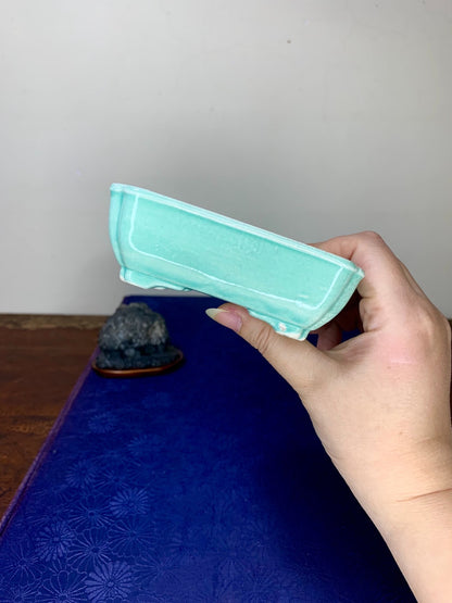 A hand holds a light turquoise glazed rectangular pot with a slightly raised rim.