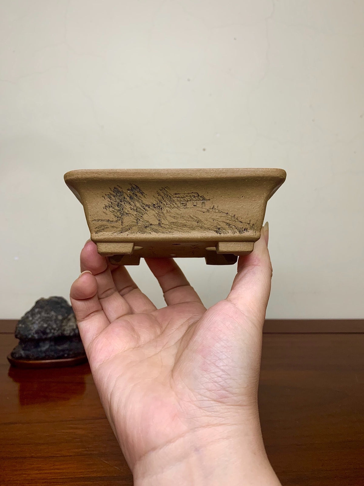 A hand holds a small, rectangular, unglazed terracotta pot with a carved mountain scene.