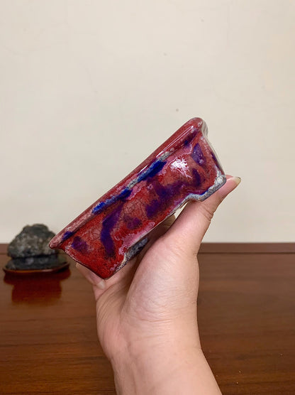 A hand holds a rectangular glazed pot with red and blue swirling glaze.